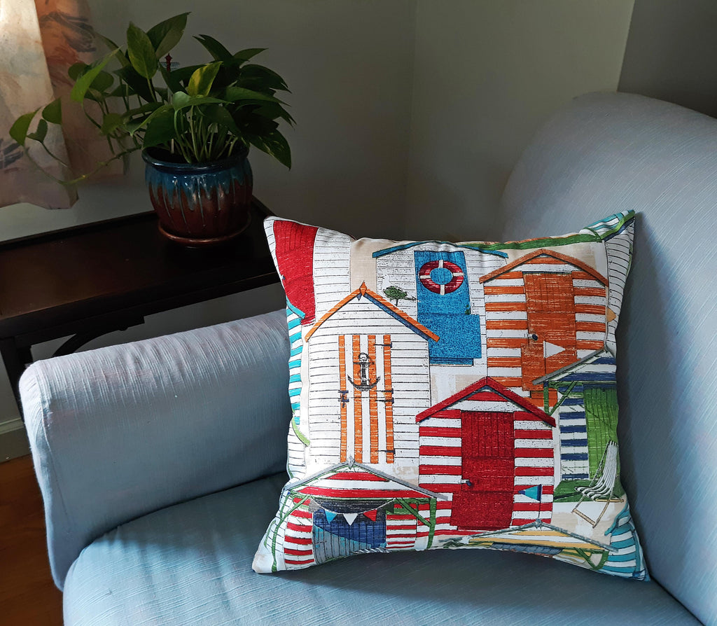 Handmade 18x18 beach hut throw pillow cover with colorful coastal cabana print in red, blue, orange and green waterproof indoor/outdoor fabric, styled on a blue armchair