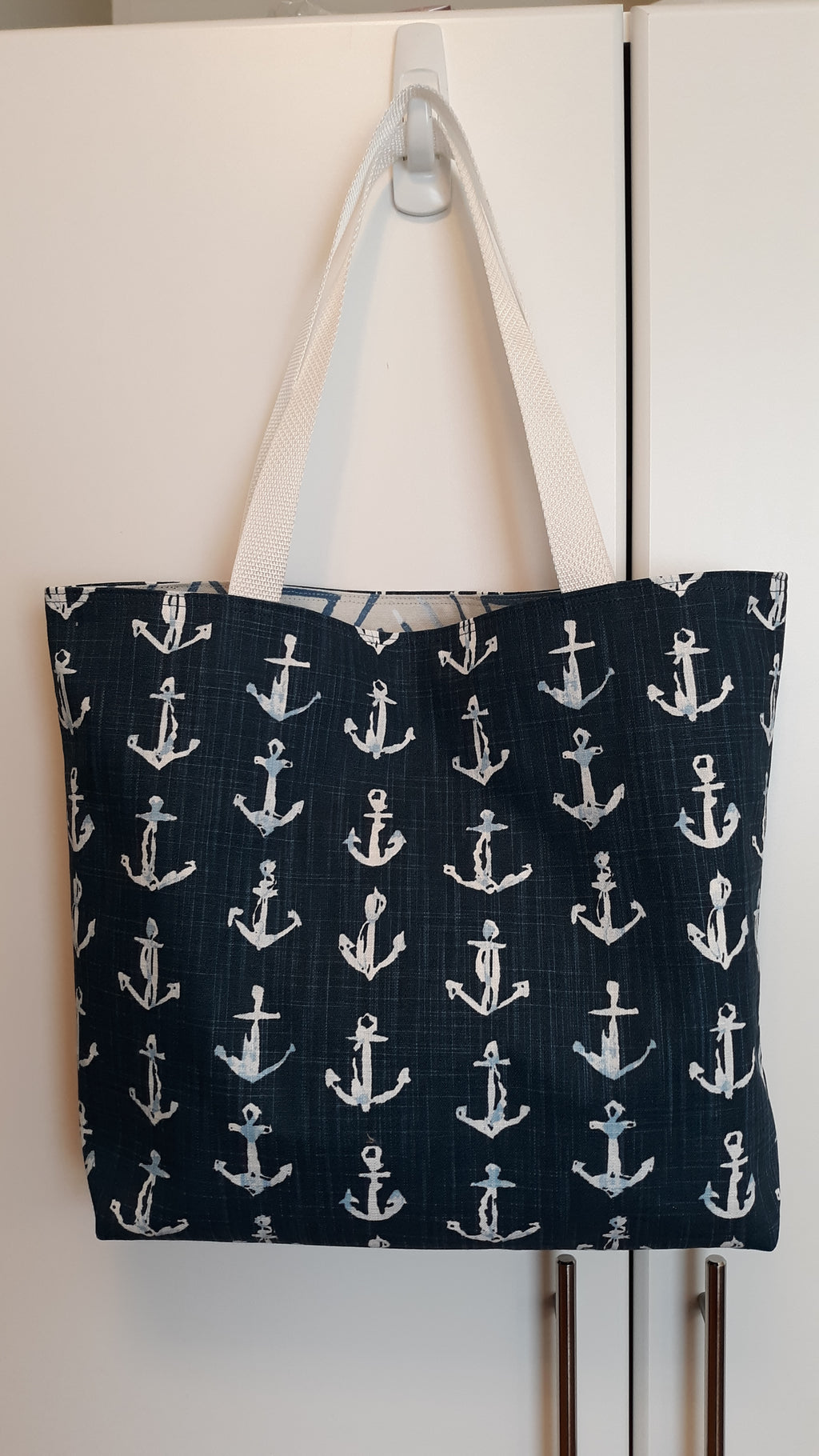 A large fabric tote bag with a navy blue background and a repeating white anchor pattern, hanging from a white plastic hook on a white door.
