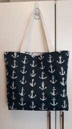 A large fabric tote bag with a navy blue background and a repeating white anchor pattern, hanging from a white plastic hook on a white door.